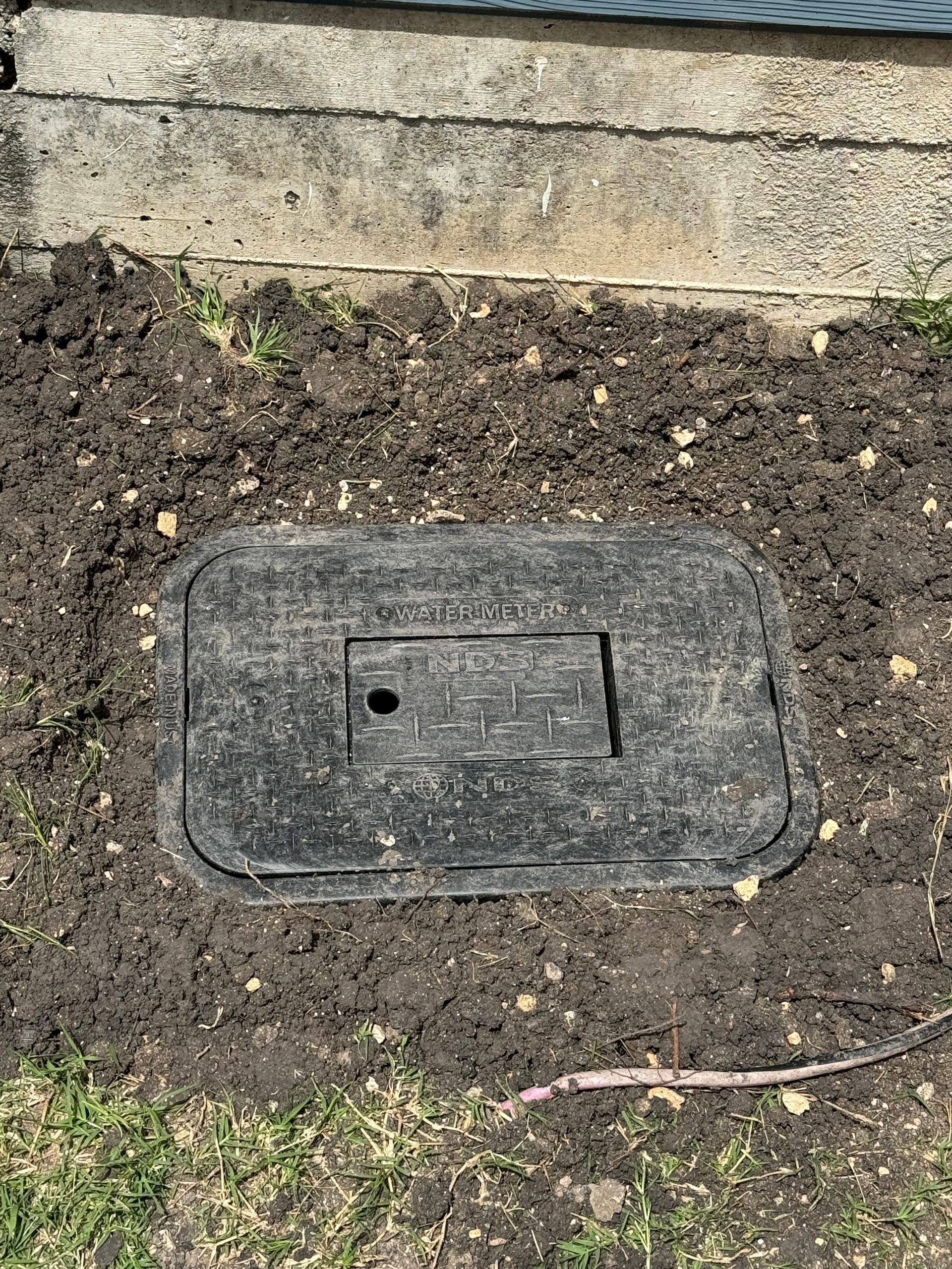 A manhole cover is sitting in the dirt next to a concrete wall.