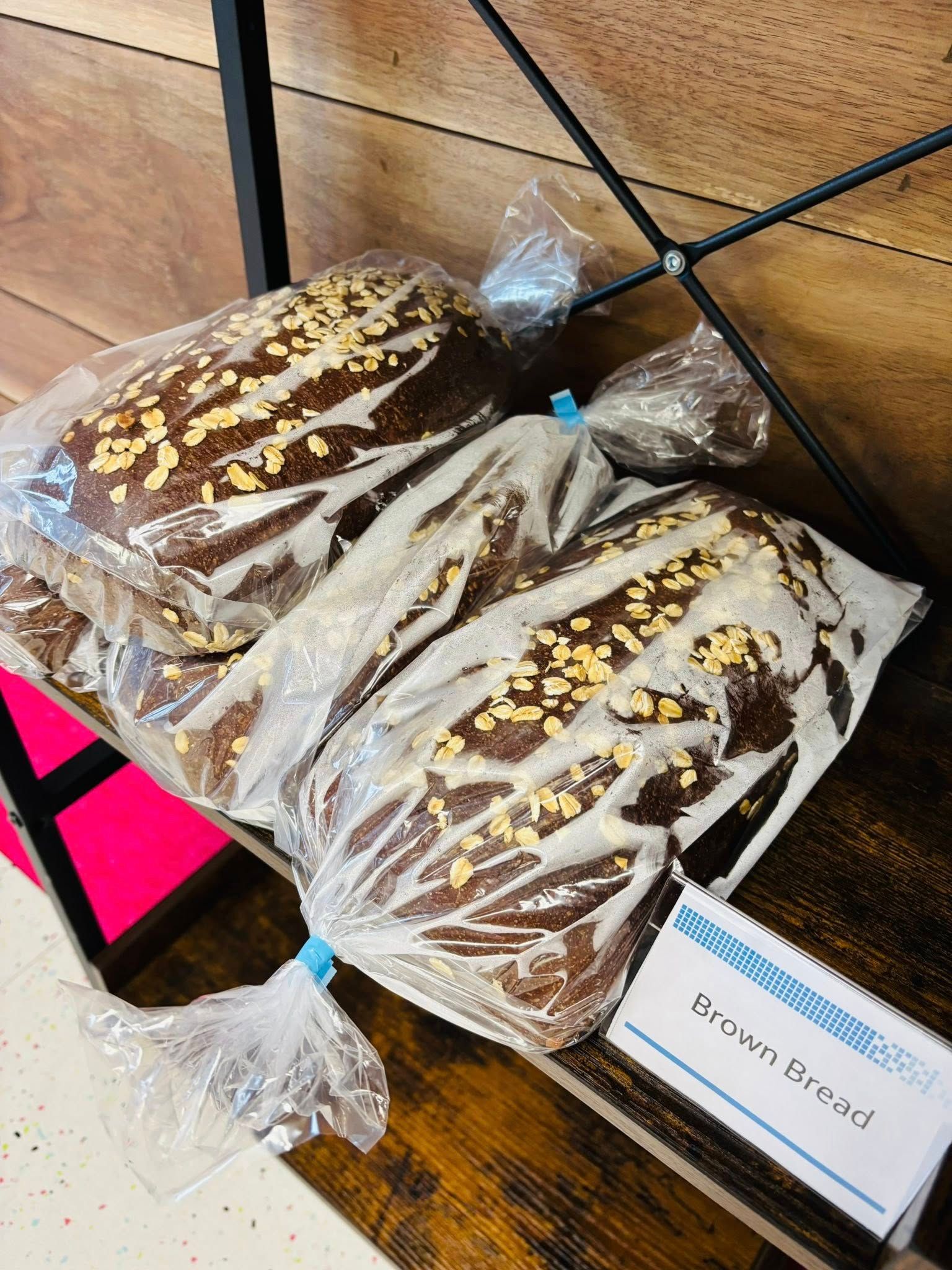 Loaves of bread in plastic bags, topped with seeds, displayed on a wooden shelf with a price tag.