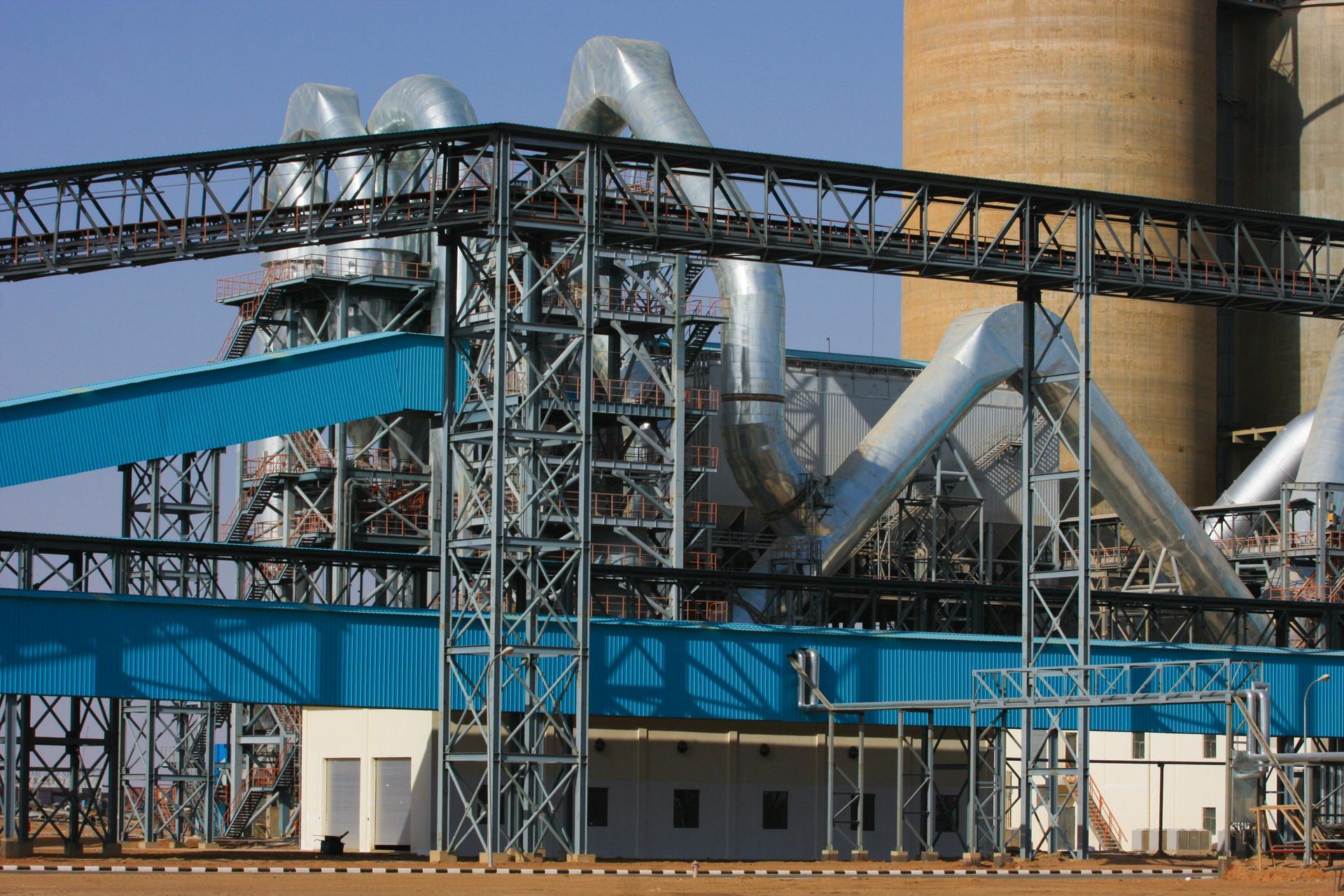 Industrial cement plant with blue walkways, metal framework, and a tall smokestack.