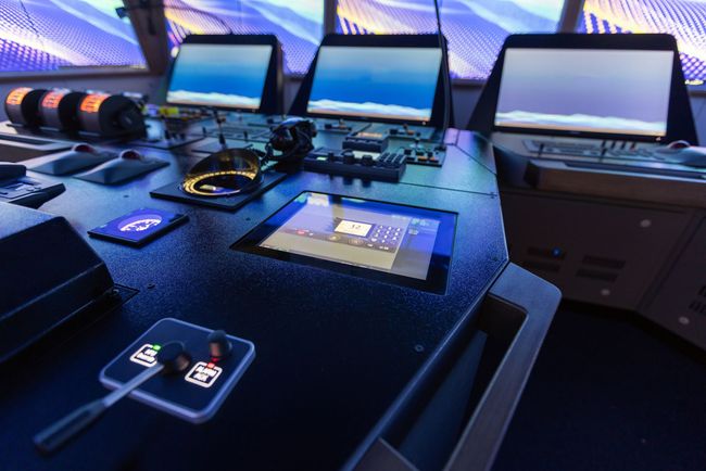 Control panel with multiple screens, buttons, and a touchscreen. Dark, futuristic setting, possibly a ship's bridge.