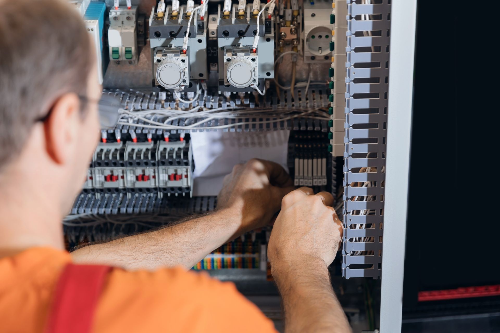 An electrician working on an electrical panel, wearing an orange shirt and overalls.