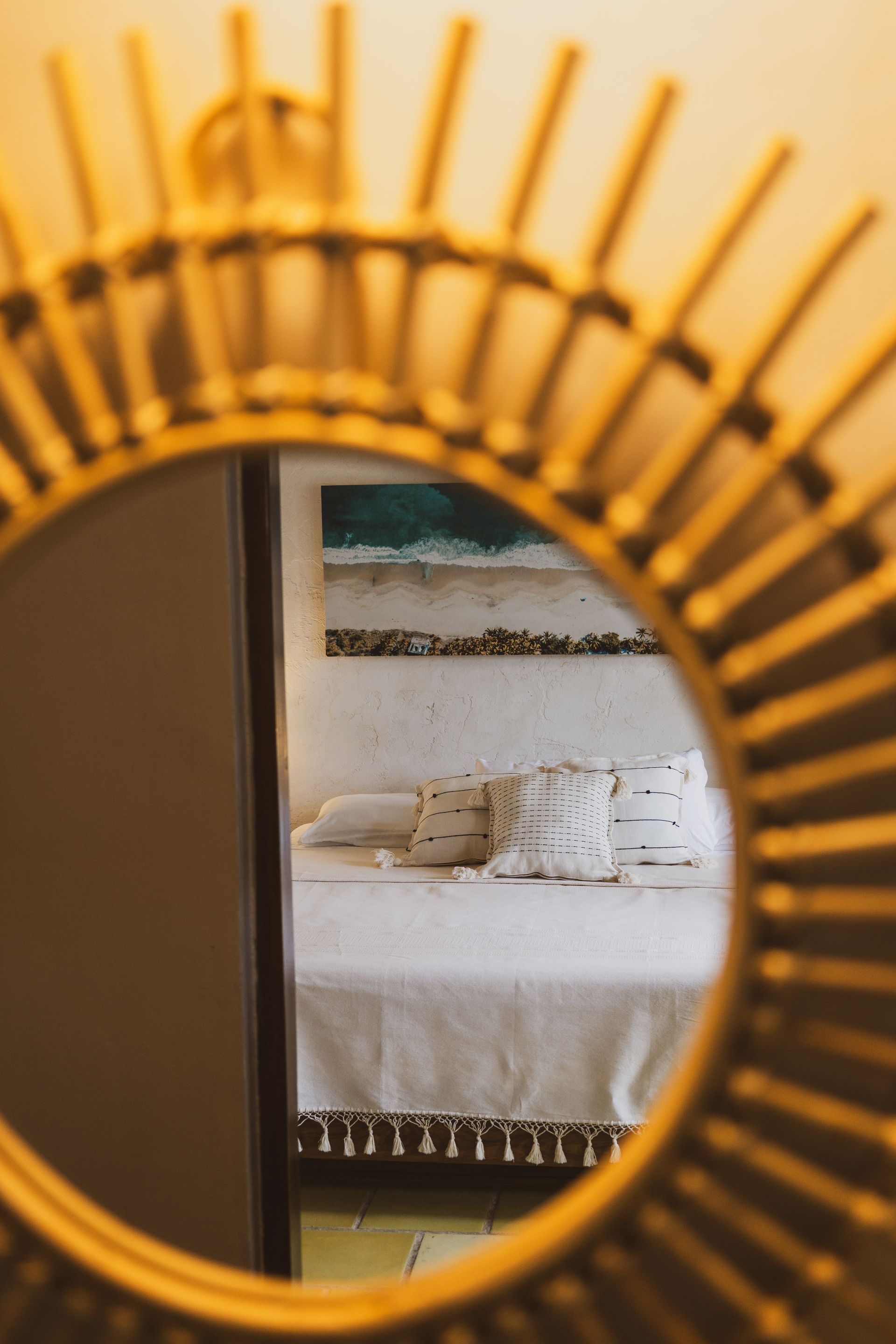 Un espejo con efecto sol refleja una cama con almohadas decorativas y obras de arte encima.