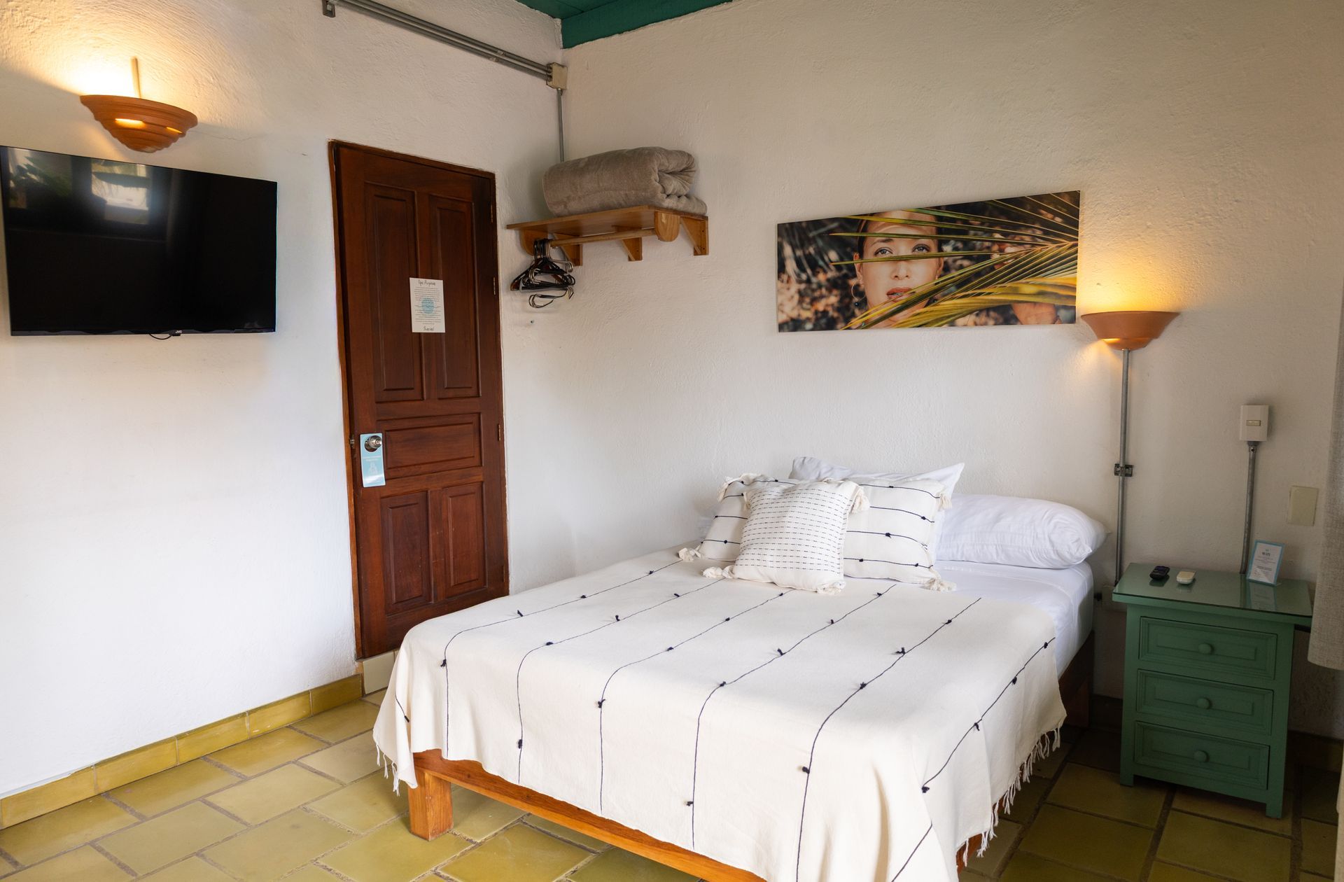 Dormitorio con cama individual, TV, puerta de madera, obras de arte y mesita de noche; paredes blancas, suelo color canela.