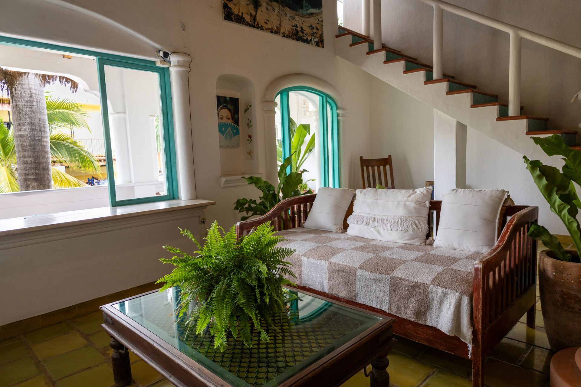 Sala de estar con sofá cama de madera, plantas, mesa de cristal y escalera. Espacio luminoso con detalles en turquesa.
