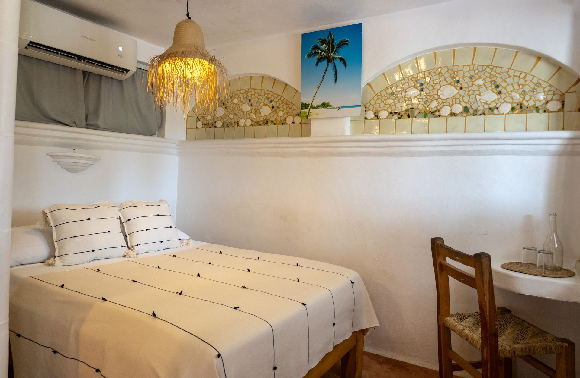 Pequeño dormitorio blanco con una cama doble, una lámpara tejida y arte mural que representa una escena de playa.