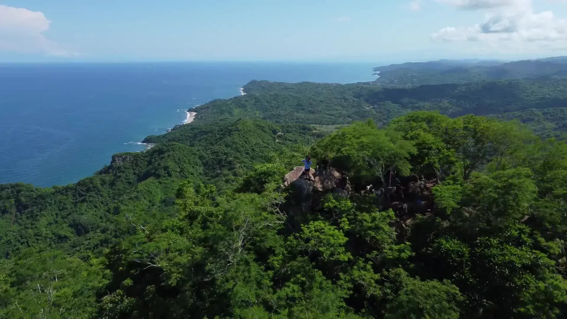 Con vistas al océano, una ladera boscosa con un pequeño edificio en la cima. Agua y cielo azules.