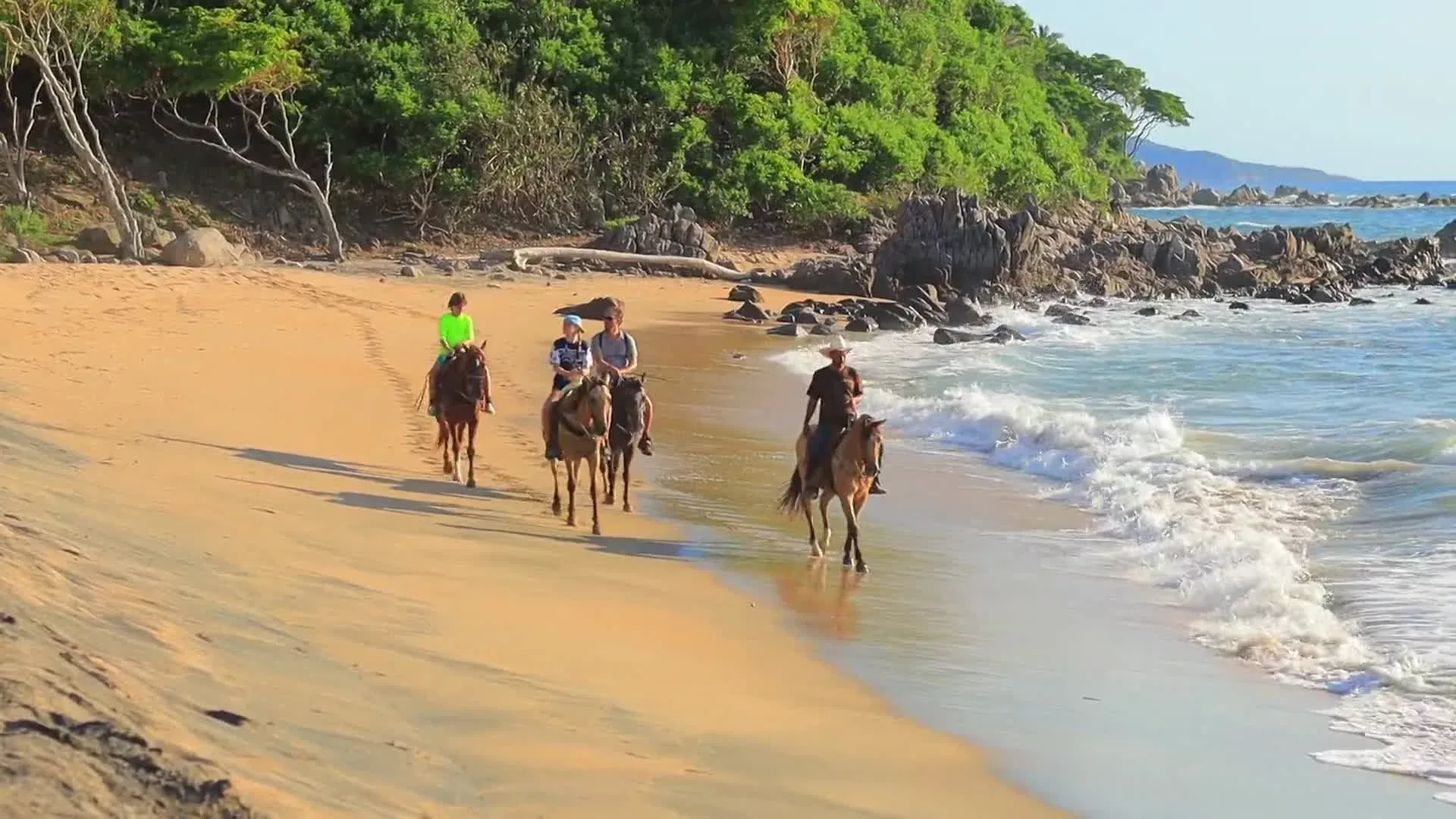 Tres personas montando a caballo por una playa de arena con olas, follaje verde y océano azul.