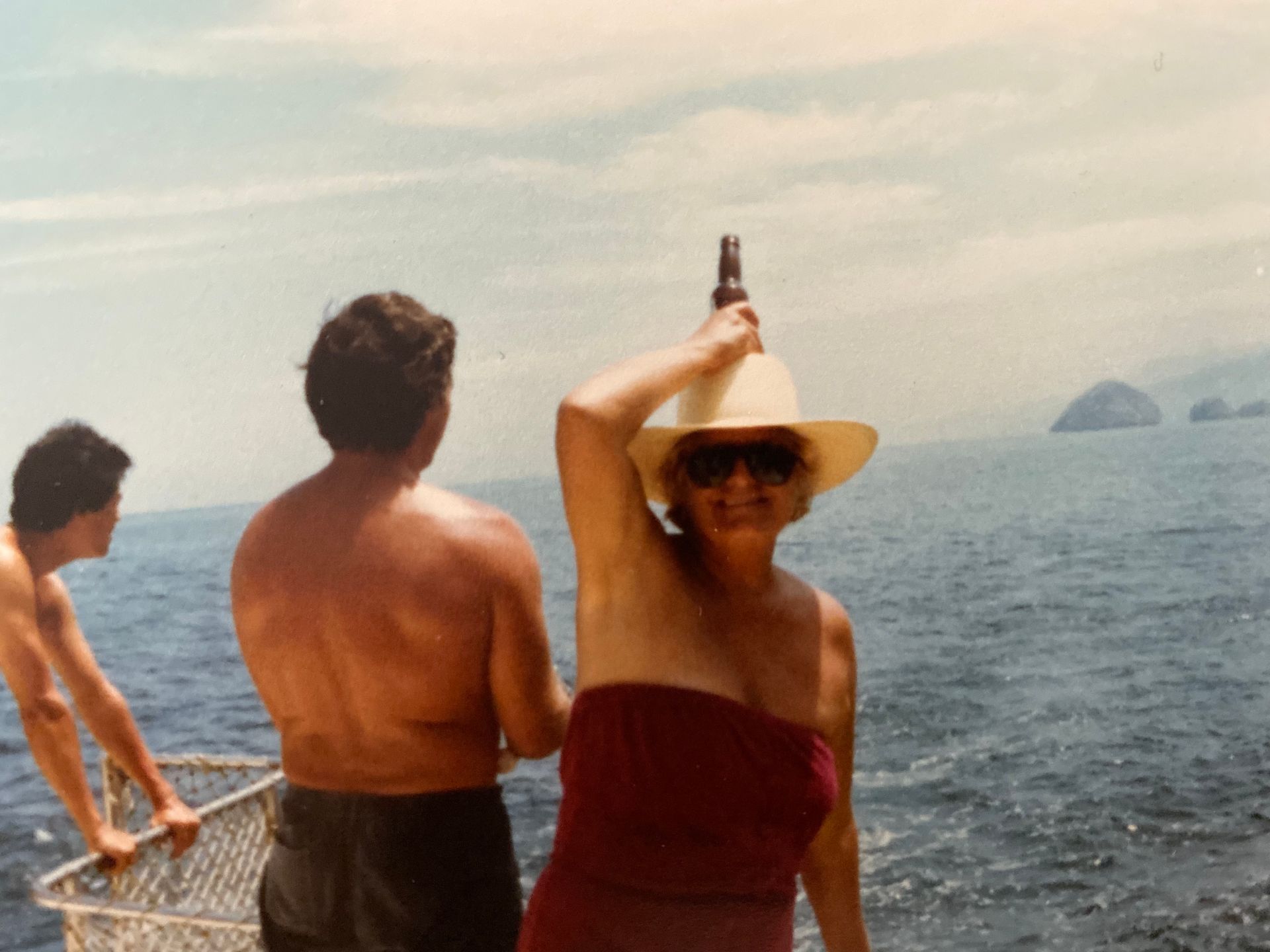 Mujer con una botella en la cabeza, de pie en un barco, con el océano de fondo. Dos hombres al fondo.