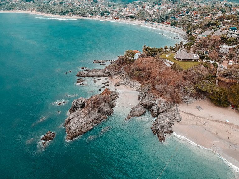 Vista aérea de una costa rocosa que se encuentra con un océano turquesa, con una playa y una ciudad al fondo.