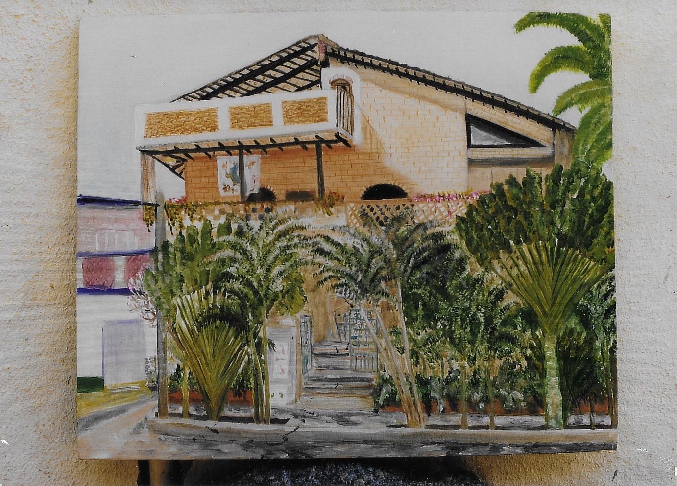 Casa de dos pisos con balcón, rodeada de plantas tropicales y calle.