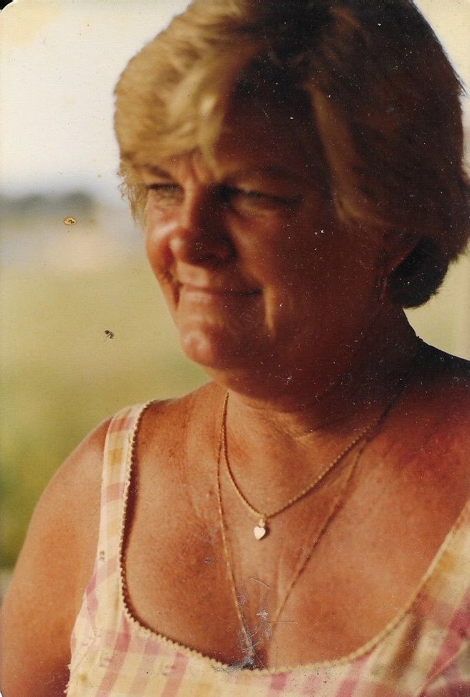 Mujer con cabello rubio corto, vestida con una camiseta a cuadros y dos collares, al aire libre.