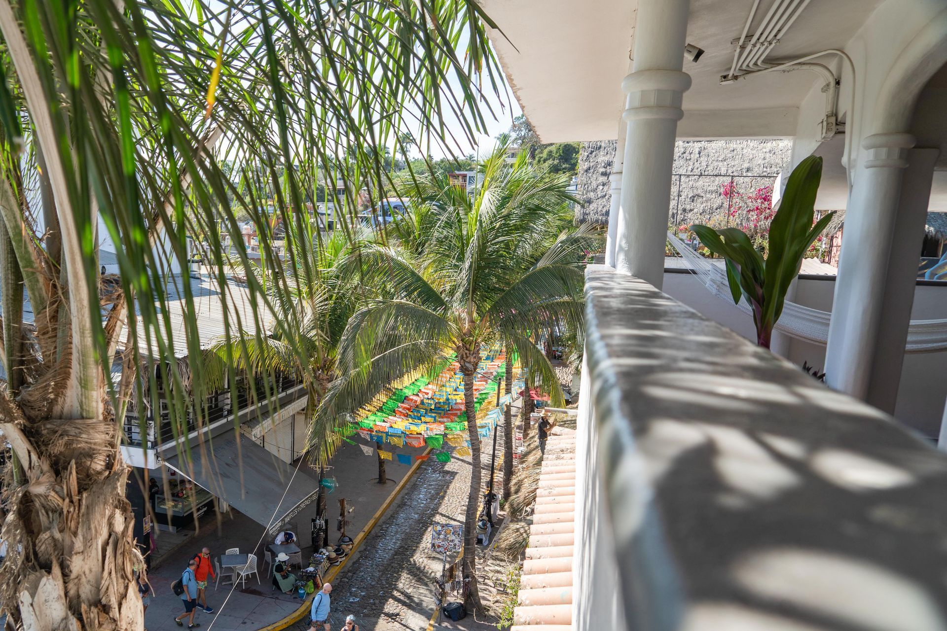 Vista desde un balcón de una calle con palmeras, tiendas y gente paseando. Luz solar brillante.