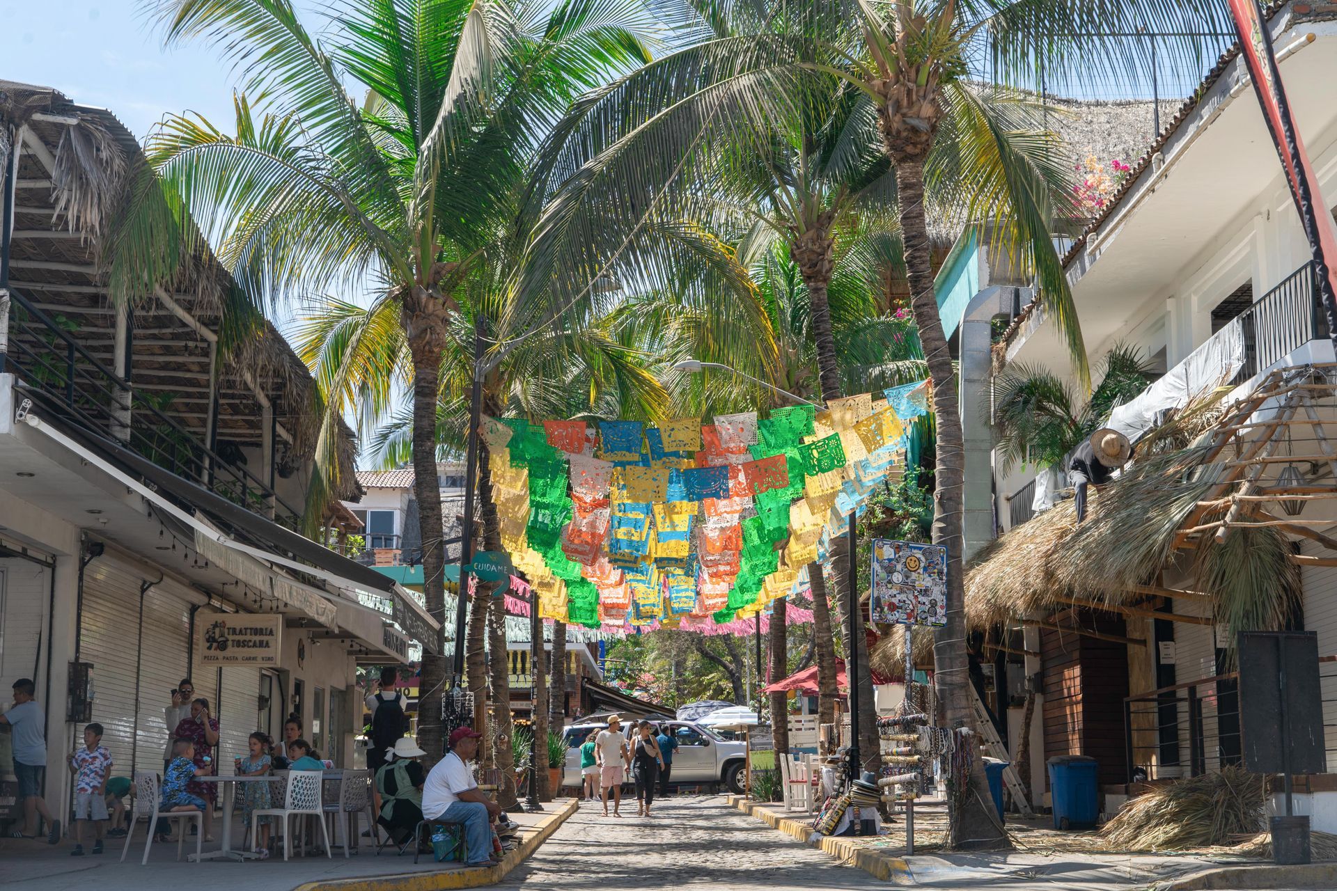 Calle llena de tiendas y palmeras, con una colorida pancarta de papel picado en el cielo. Gente paseando y sentada afuera.