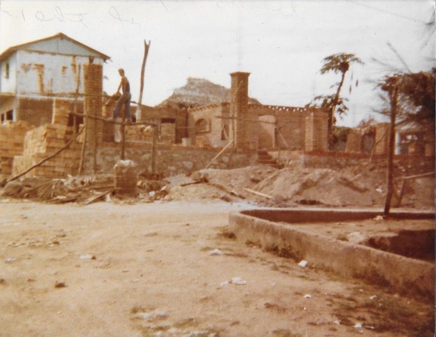 Ruinas de un edificio de ladrillo demolido, suelo de tierra, persona de pie, casa azul al fondo.