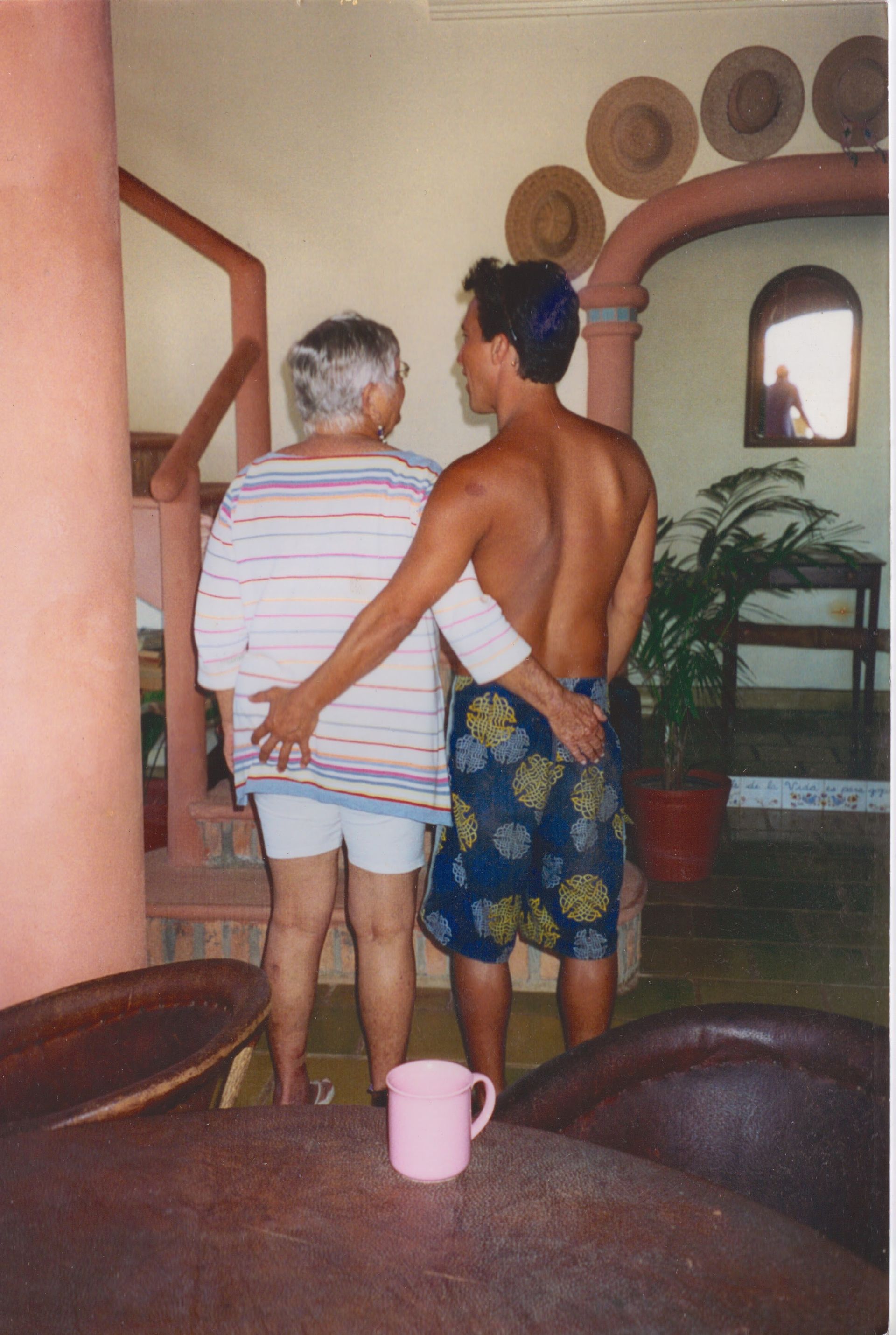 Un hombre rodea a una persona con el brazo; la toca por detrás. Una taza rosa reposa en primer plano. Escenario interior.