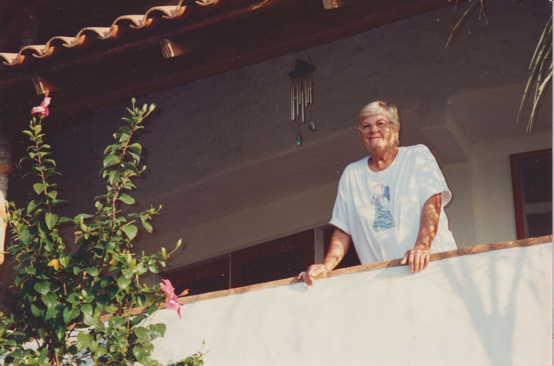 Mujer apoyada en un balcón, sonriendo. Lleva una camisa blanca; follaje en primer plano.