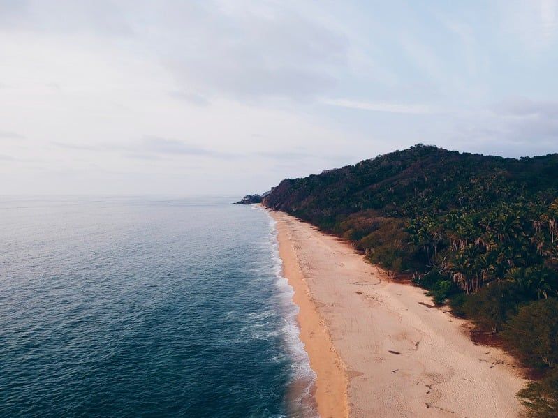 Olas del océano rompiendo en una playa de arena, bordeada por una exuberante y verde ladera bajo un cielo nublado.