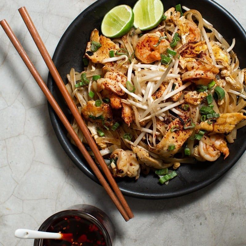Pad Thai con camarones y pollo en un tazón negro, palillos, limón, aceite de chile.
