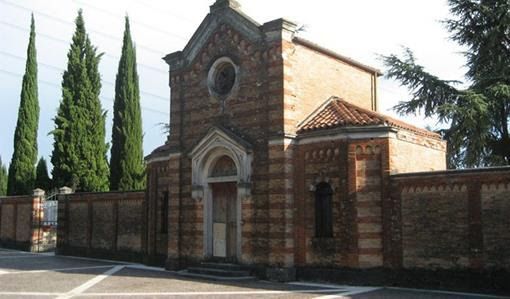 Cappella in mattoni con ingresso ad arco, piccola finestra rotonda e alti cipressi lungo un muro di mattoni.