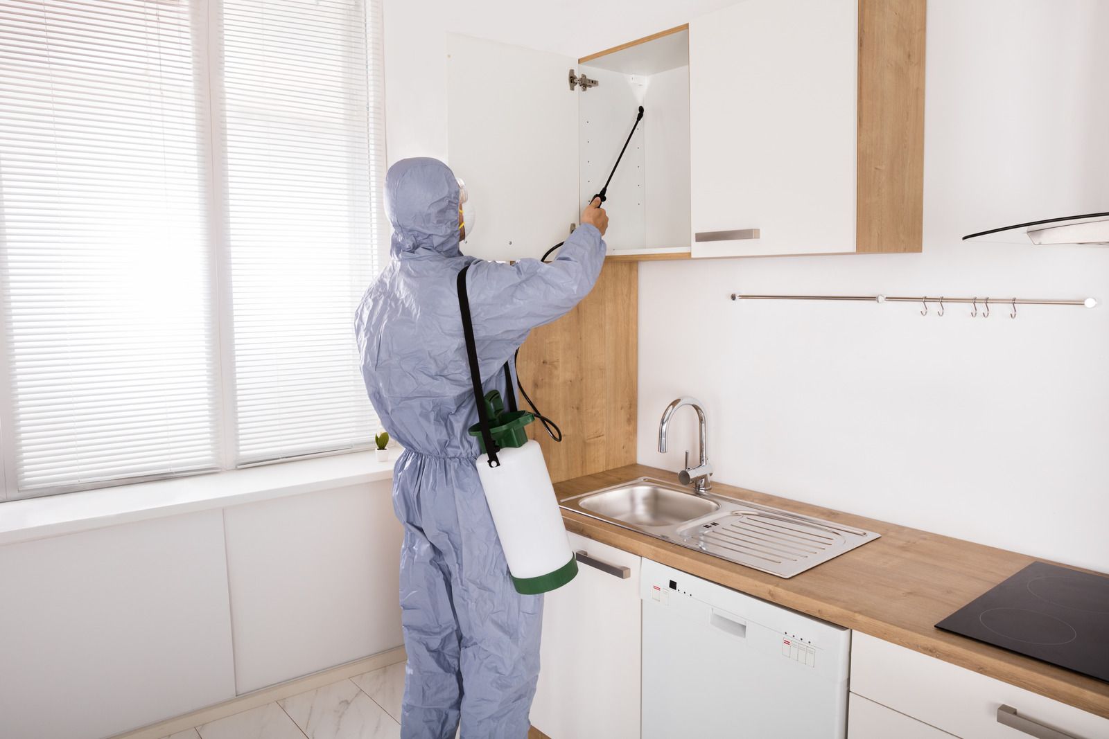 A man in a protective suit is spraying a kitchen cabinet with a sprayer.