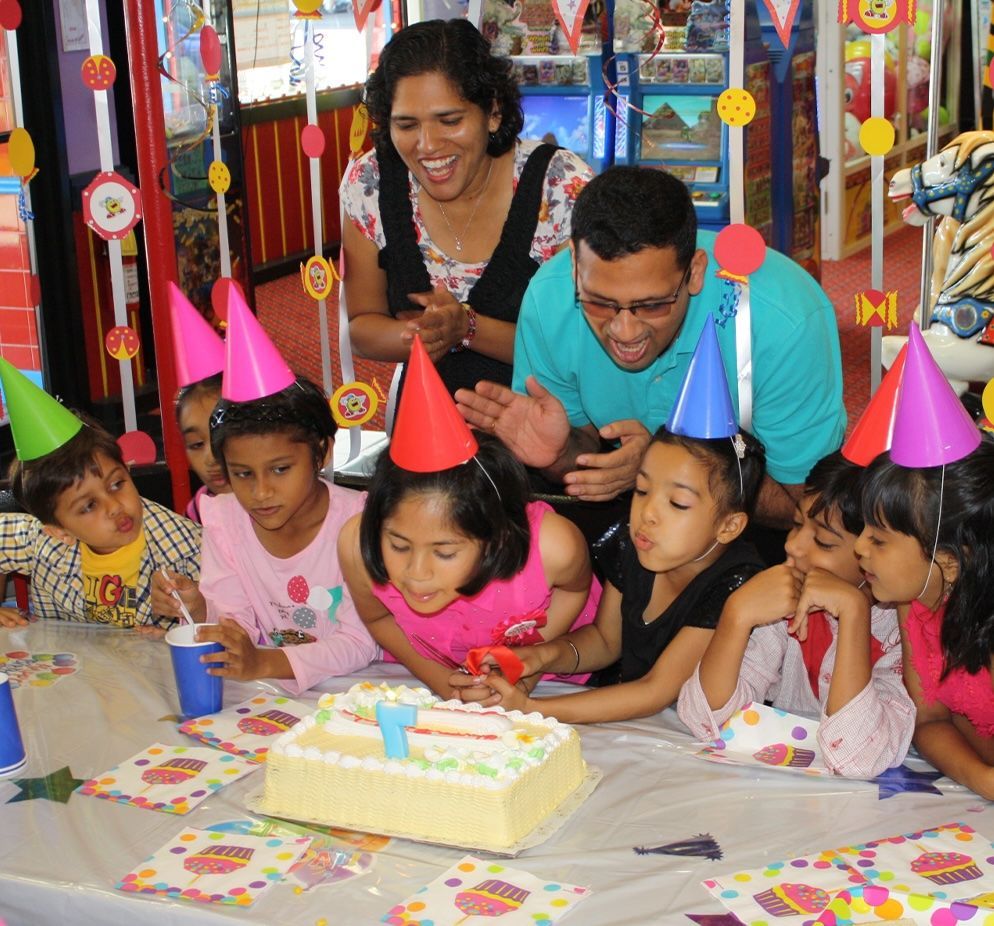 A group of children wearing party hats are gathered around a birthday cake