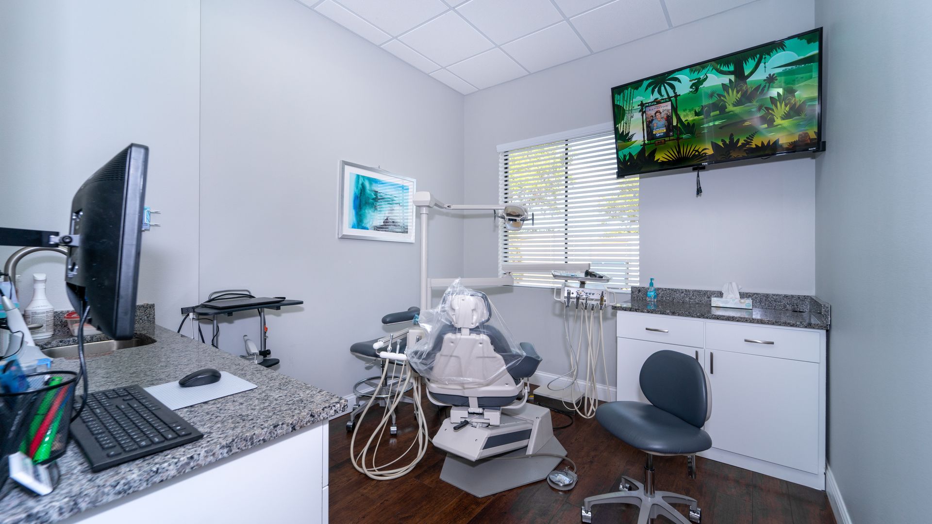 A dental office with a dental chair , desk , computer and television.