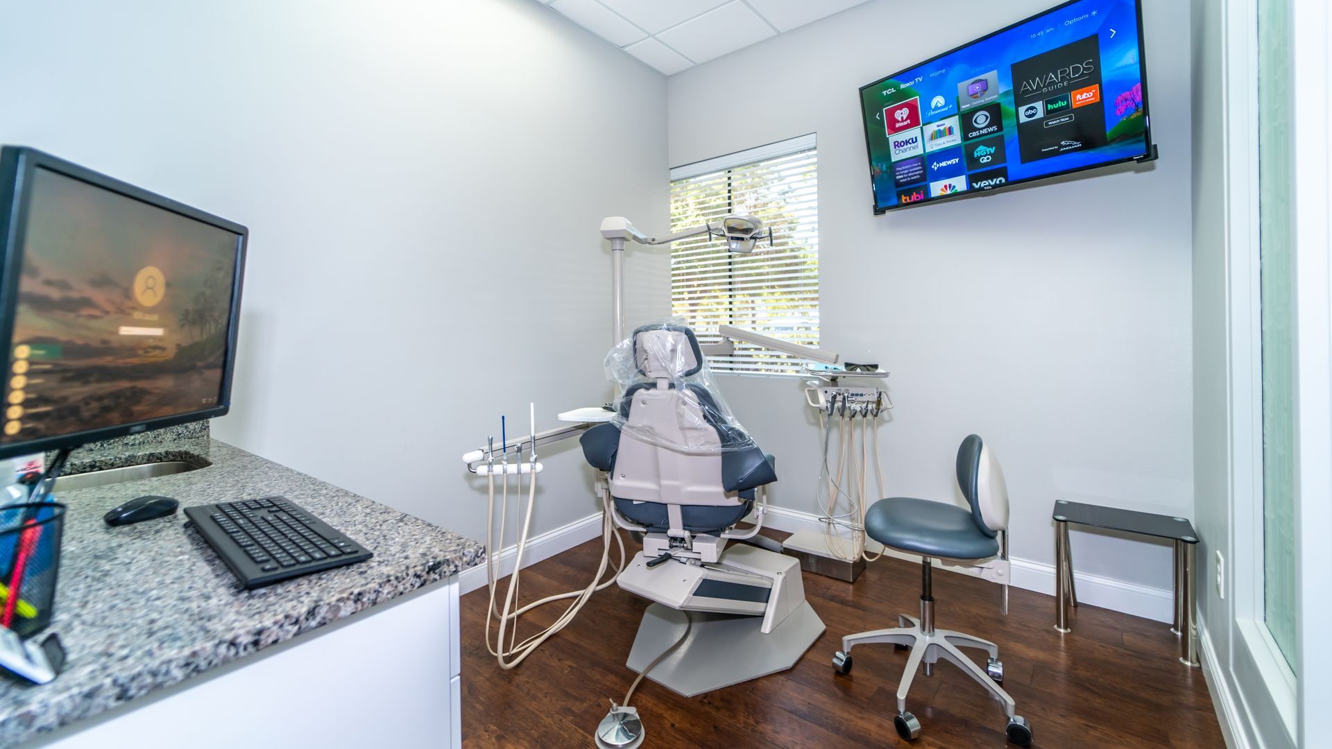 A dental office with a dental chair , desk , computer and television.