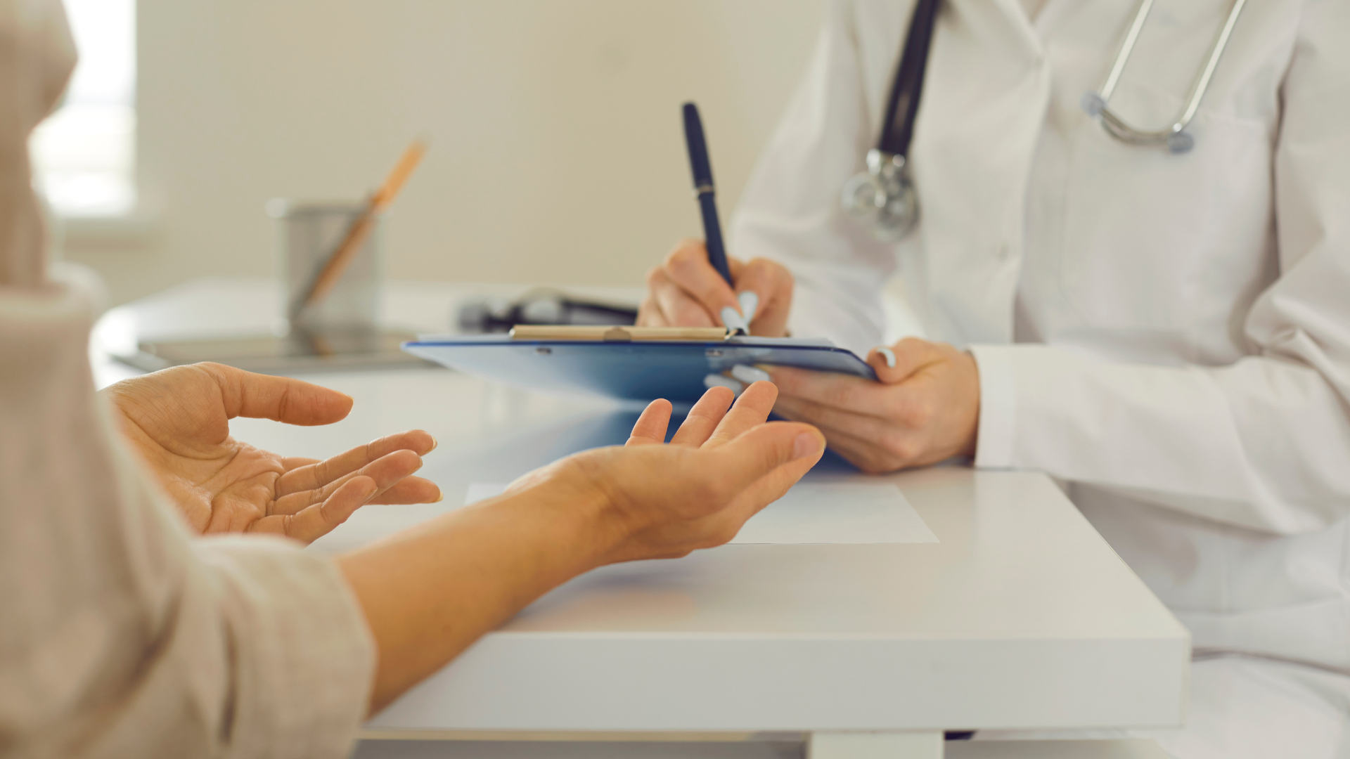 Patient at doctor's appointment, gesturing with hands. Doctor writes on clipboard in office setting.
