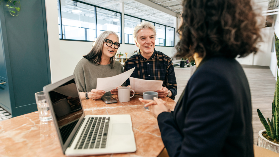 A financial advisor speaks with a couple in a bright office, reviewing papers and using a laptop.