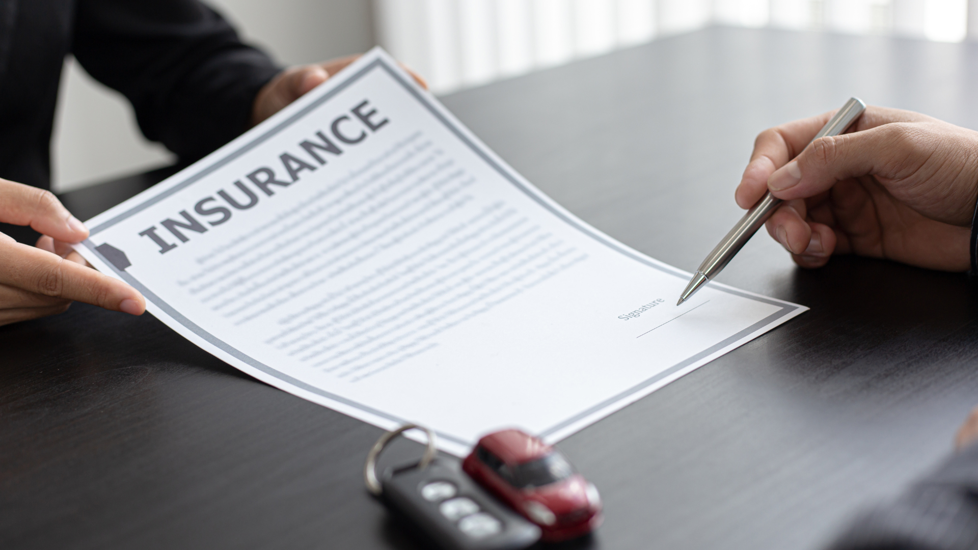 Person signing an insurance document, car keys and a toy car on a table.
