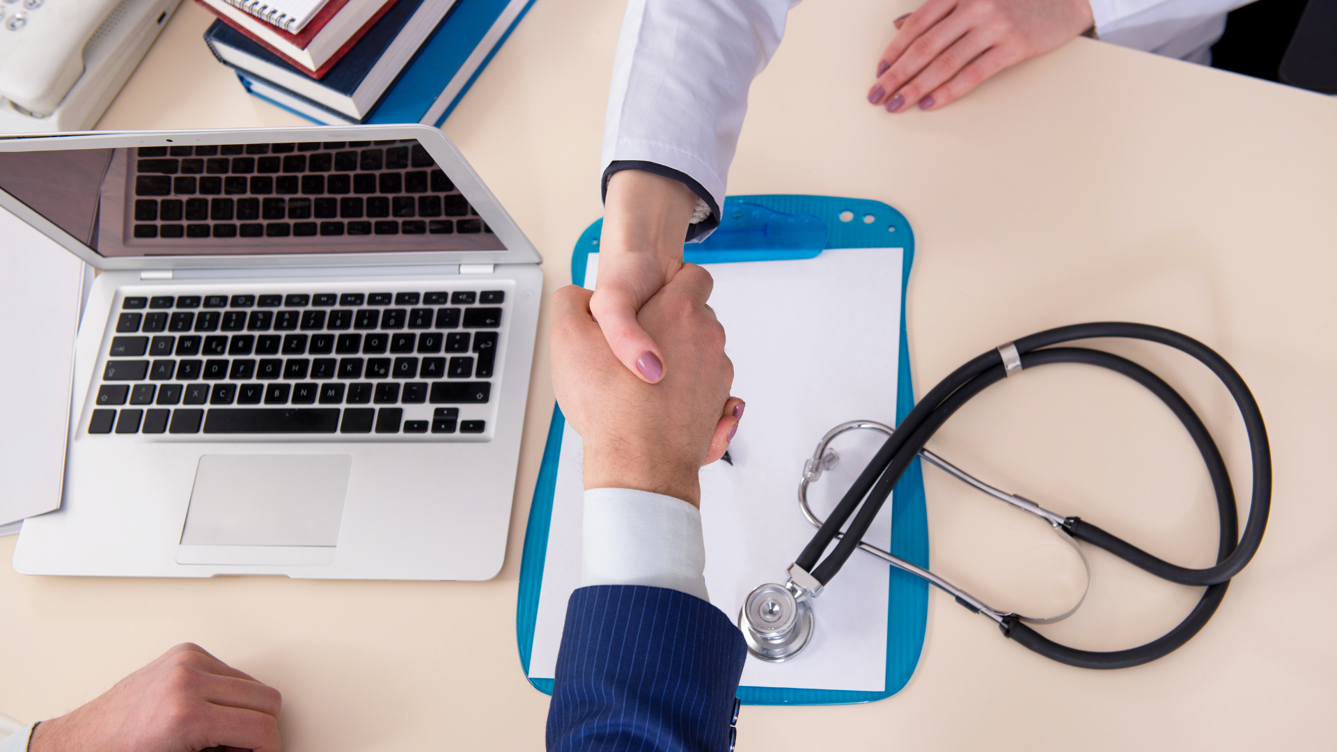 Doctor and patient shaking hands at desk with stethoscope, clipboard, and laptop.