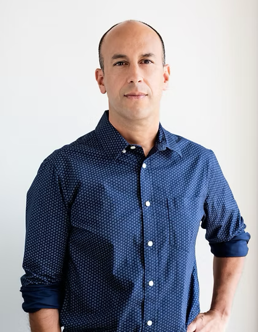 Bald man in blue button-down shirt, hand in pocket, looking at the camera. White background.