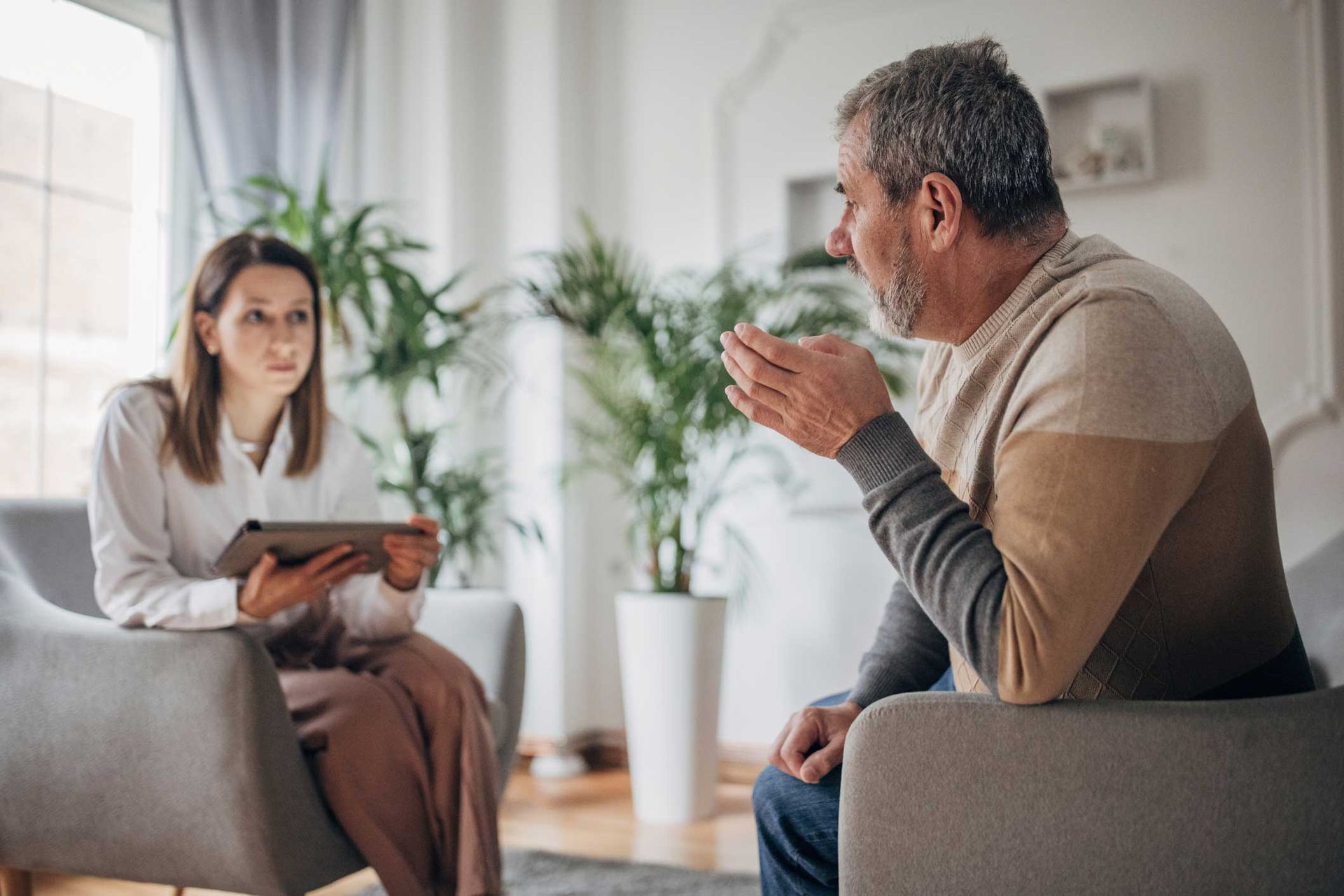 A man is sitting in a chair talking to a woman who is holding a tablet.