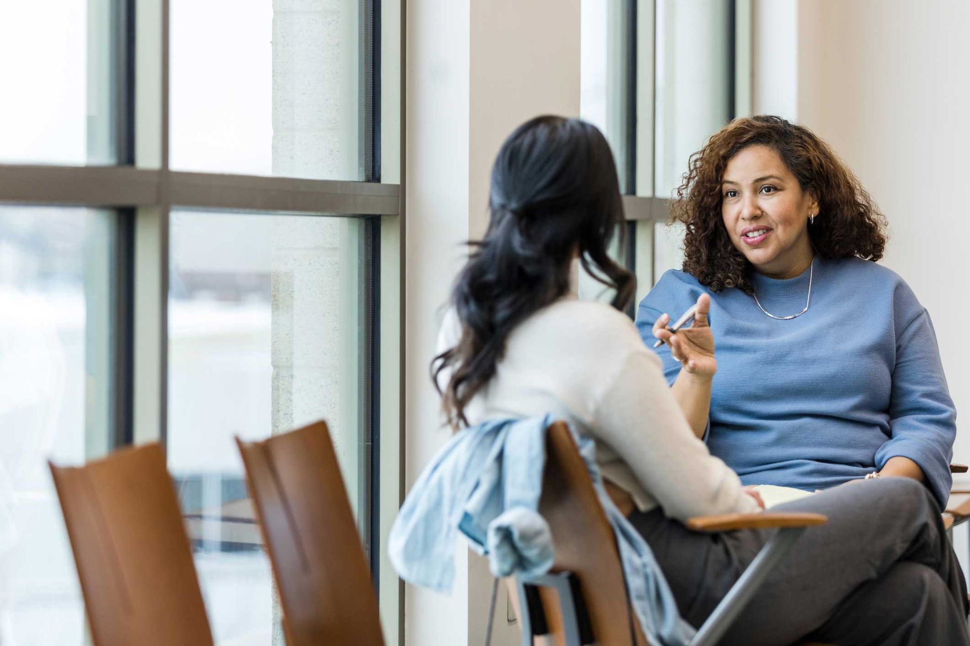 Two women are sitting in chairs talking to each other in front of a window.