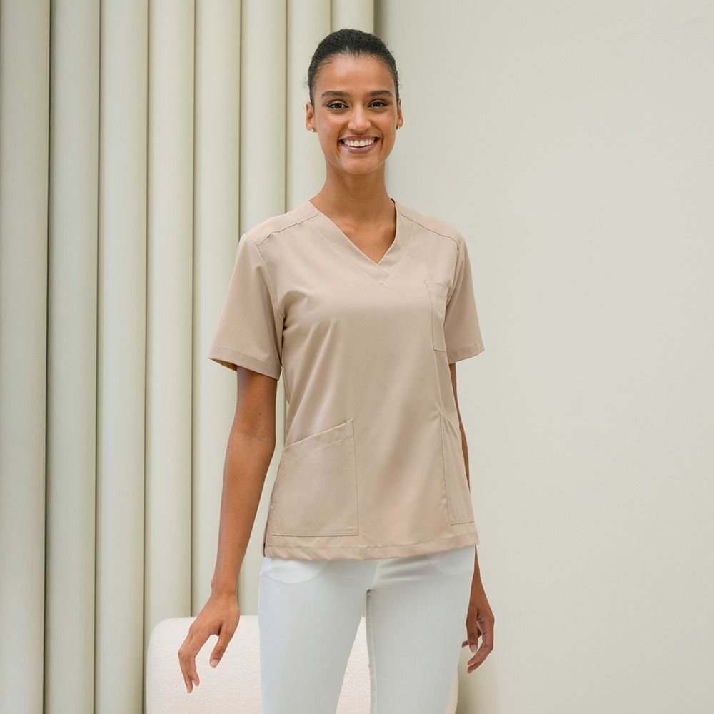 Donna sorridente che indossa una camicetta beige e pantaloni bianchi, in piedi vicino a una tenda.