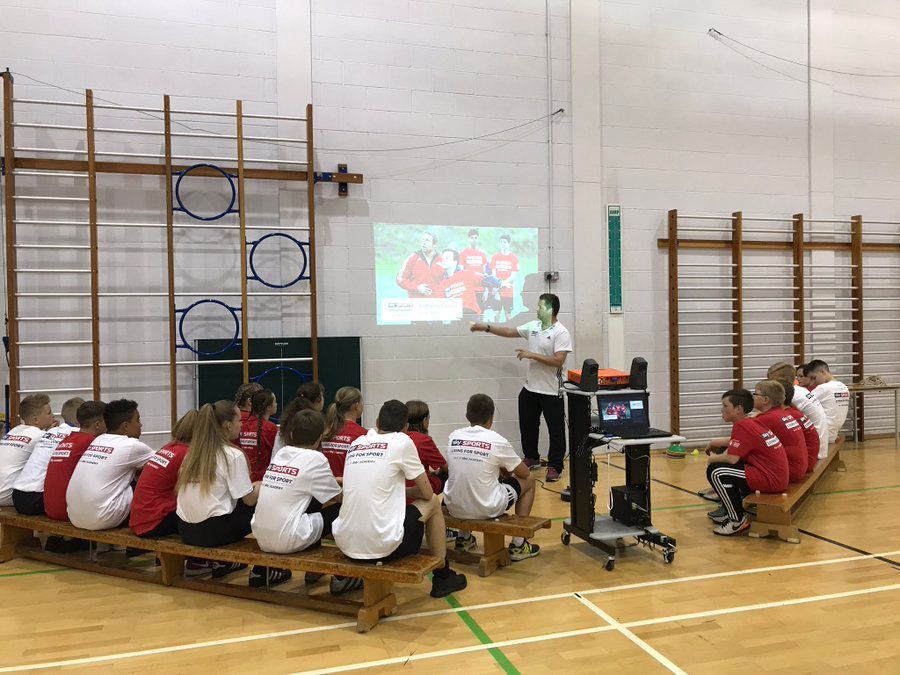 A group of people are sitting on benches in a gym watching a presentation.