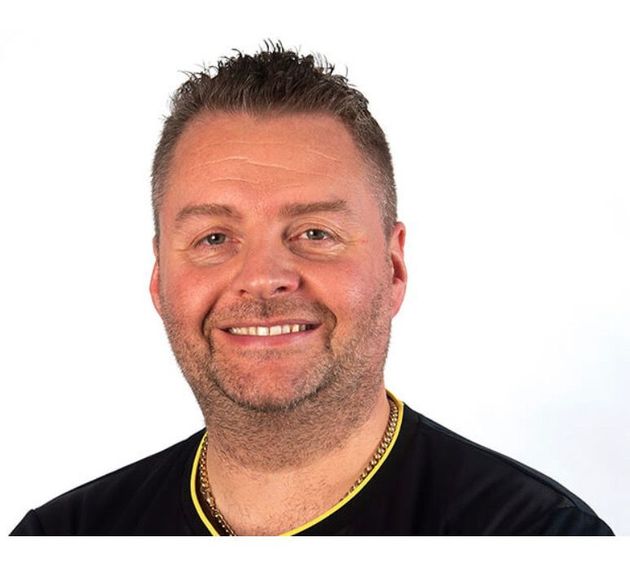 A man wearing a black shirt and a yellow chain around his neck smiles for the camera
