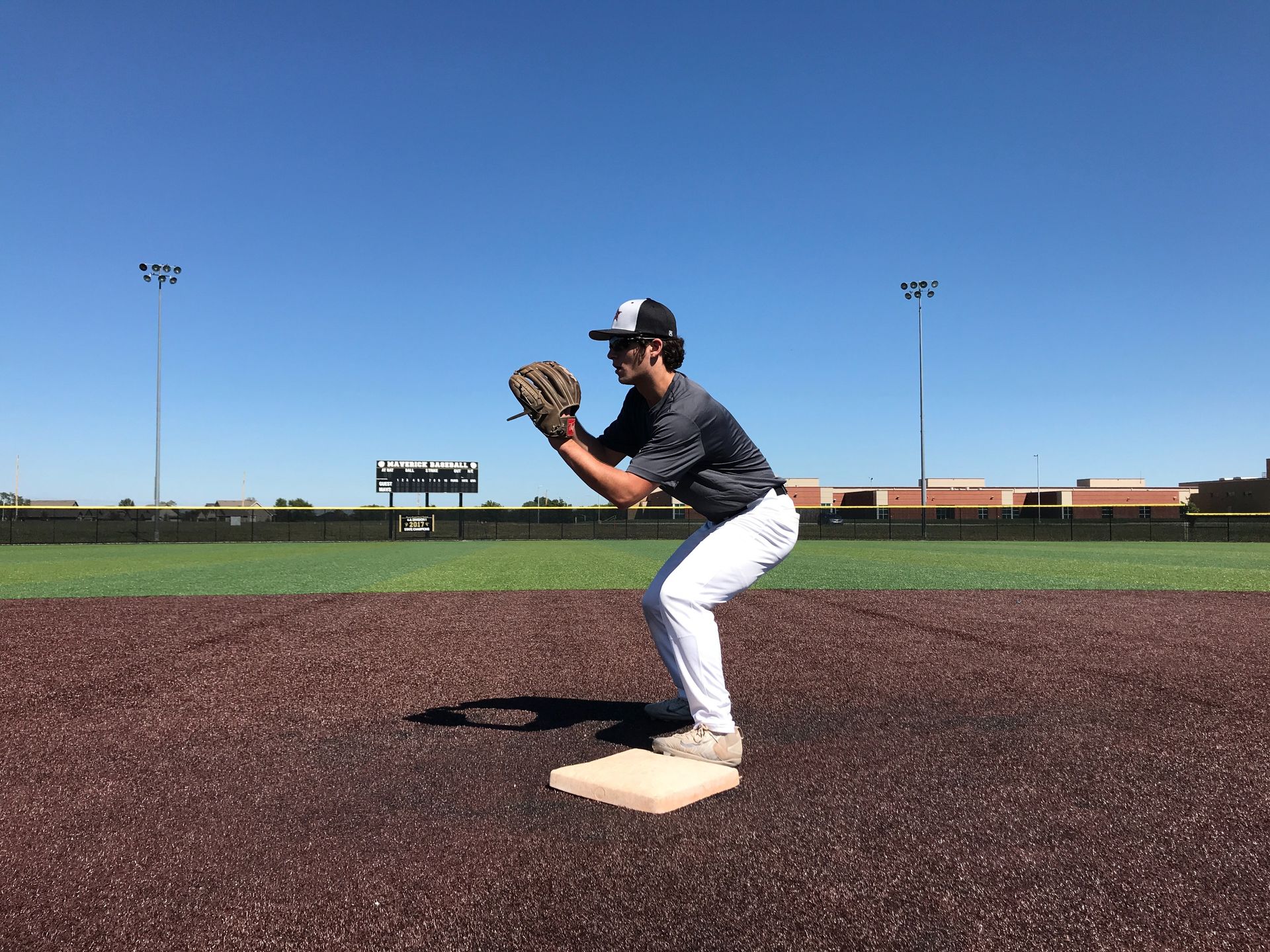 Baseball player in gray uniform crouching on a field, ready to catch a ball under a clear blue sky.