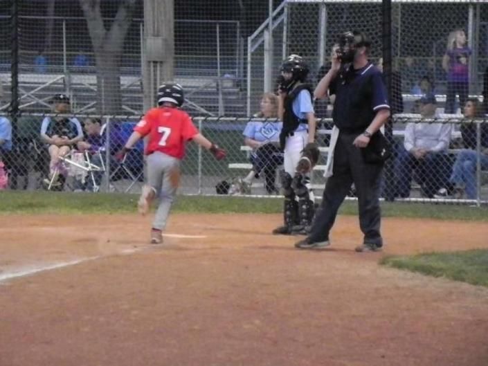 Youth baseball player in red jersey runs toward home plate as umpire and catcher watch.