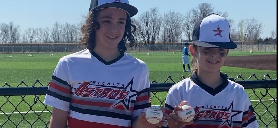 Two baseball players in black-and-red jerseys smiling on a field, holding baseballs under a blue sky