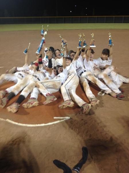Youth baseball team in white uniforms celebrating on the field with trophies raised high