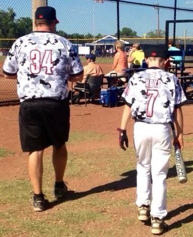 Two baseball players in white camo jerseys walk toward the dugout at a youth field.