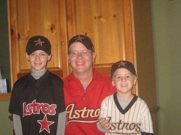 Three people in Houston Astros gear smiling indoors in front of a wooden wall