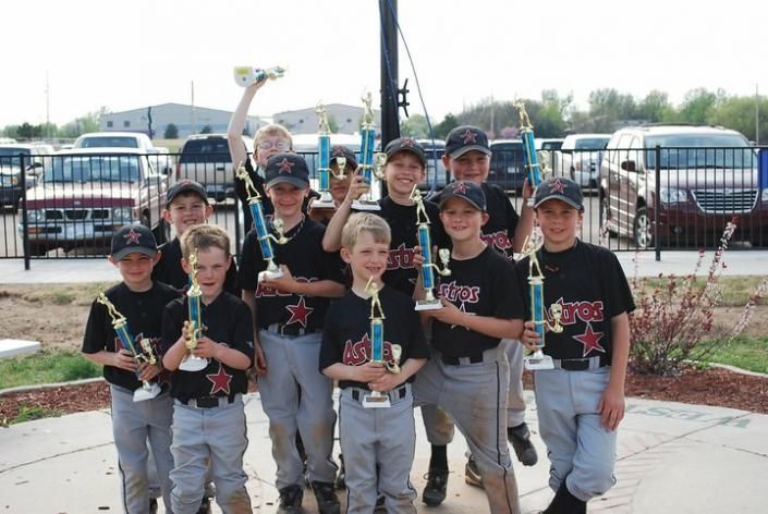 Youth baseball team holding trophies in front of parked buses, smiling outdoors.