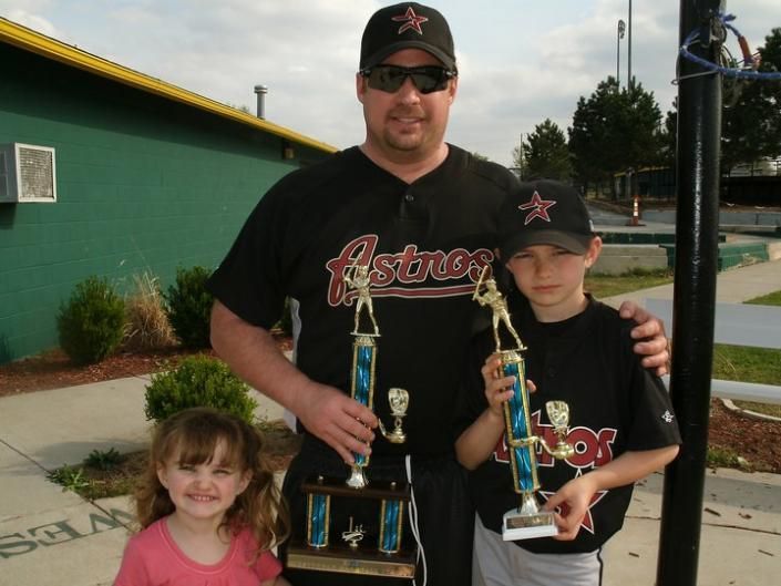 Man holding two trophies with two children outdoors in front of a building