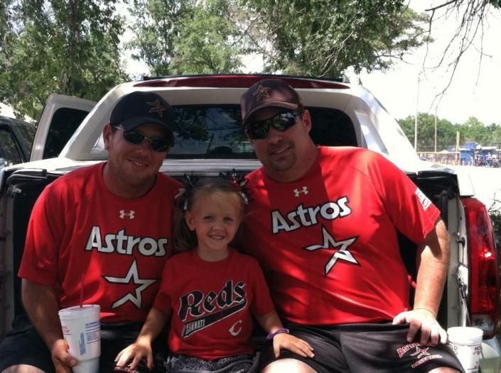 Three people in red Astros shirts smile sitting in the back of a pickup truck outdoors