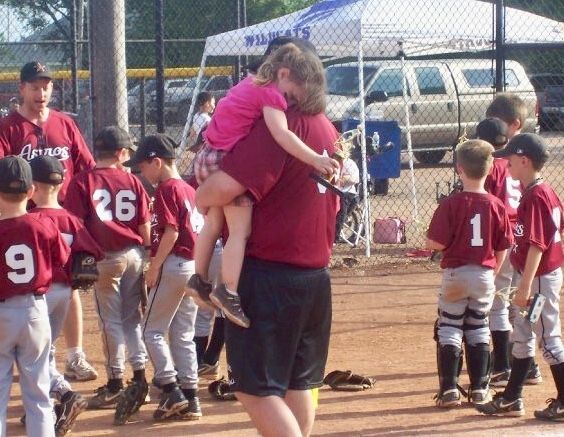 Youth baseball team gathered near a coach holding a child, with players in maroon jerseys by the dugout.