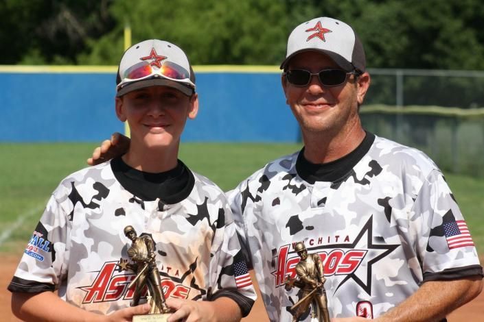 Two baseball players in camouflage jerseys and caps posing with trophies on a field