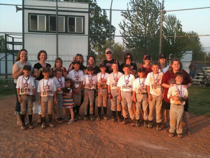 Group of children and adults standing together on a dirt court at sunset, wearing white shirts and tan pants.