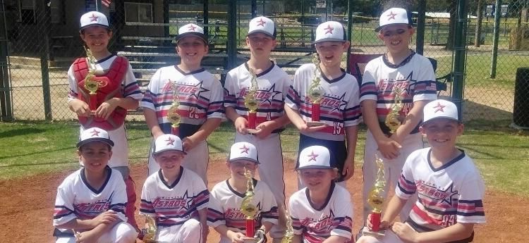 Youth baseball team posing in uniforms on a sunny field with a tree and building in the background